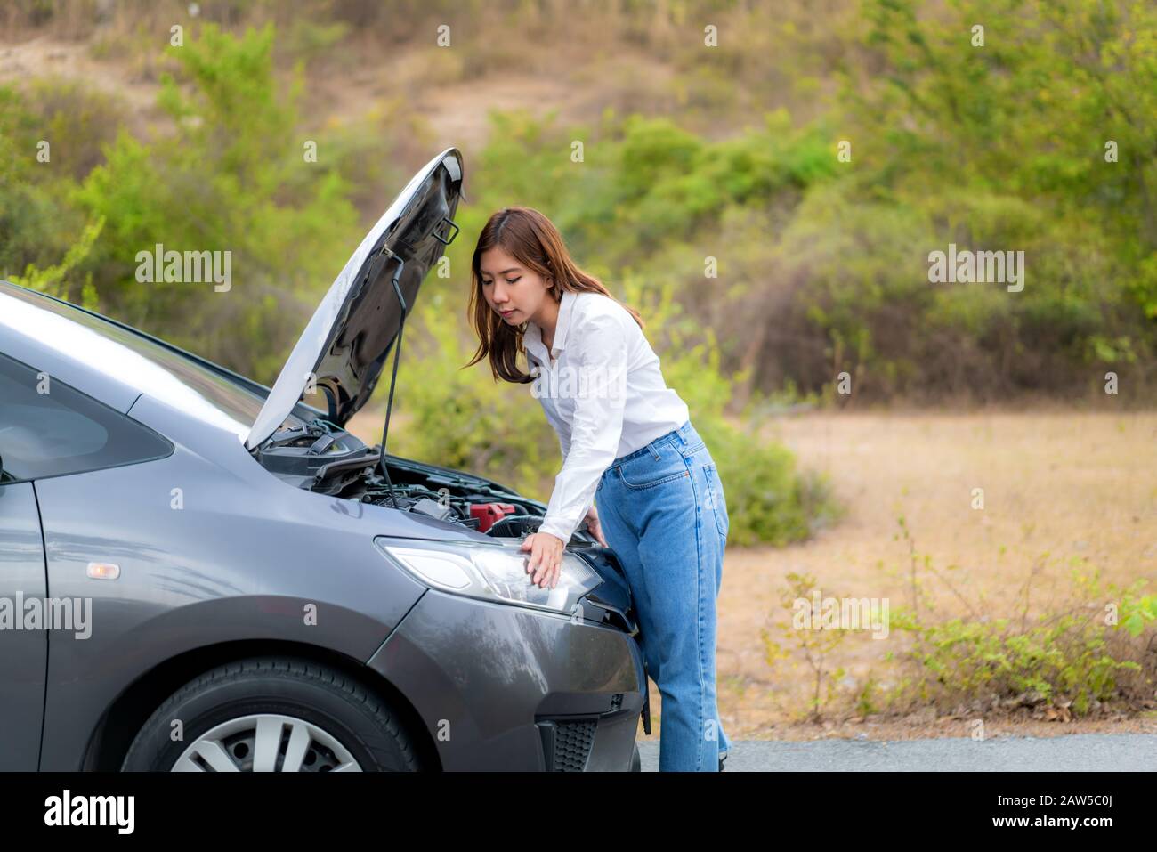 Junge, unglückliche asiatische Frau, die einen zerbrochenen Automotor vor der offenen Haube inspiziert, zerbrochenes Auto Auf Der Country Road, Das Auf die Fahrhilfe wartet Stockfoto