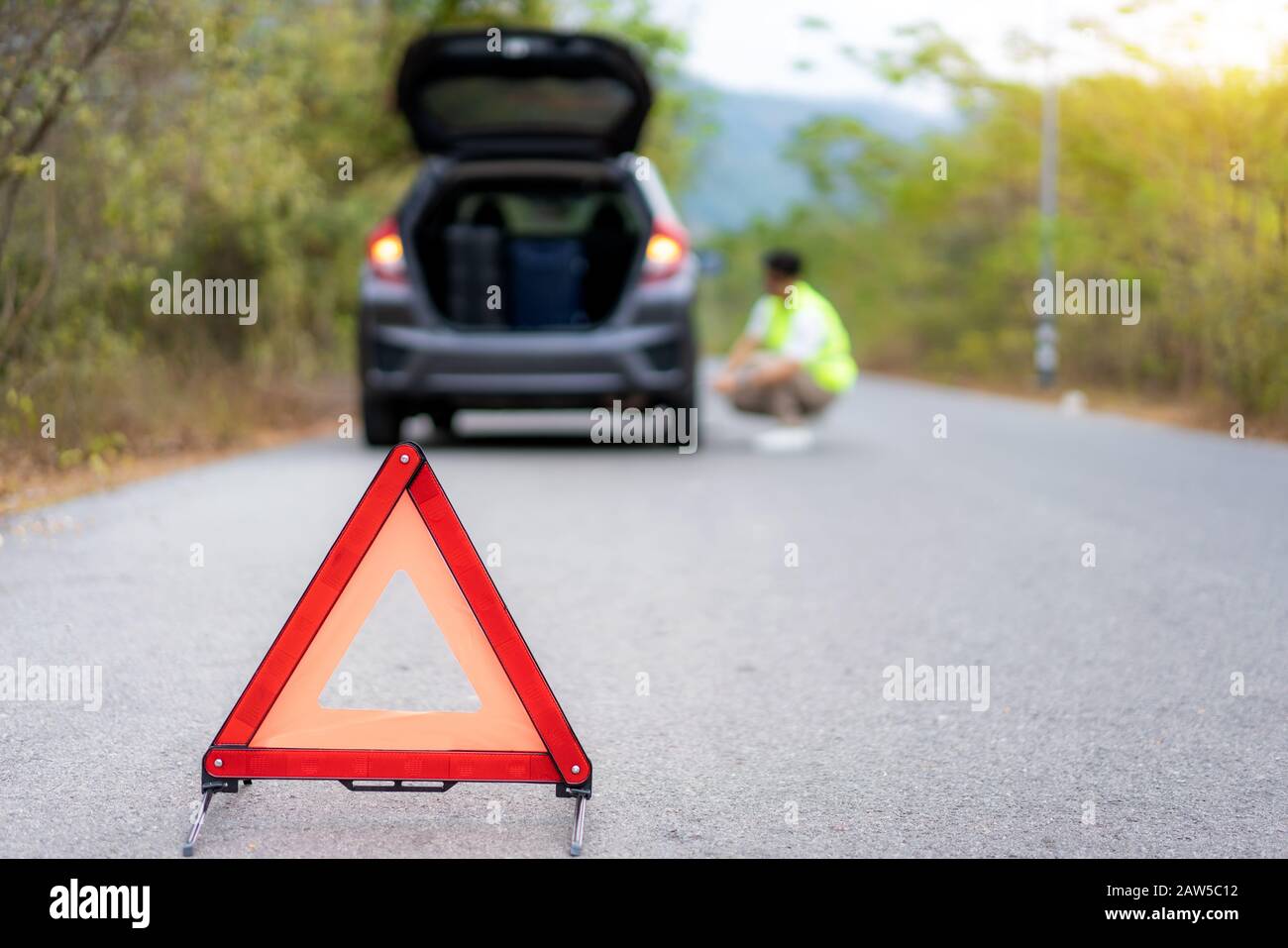 Schild mit dem Dreieck auf der Straße mit einem besorgten asiatischen Mann, der den Reifen repariert und wechselt, während die Versicherung wartet oder die Autofirma nach dem Auto kommt Stockfoto