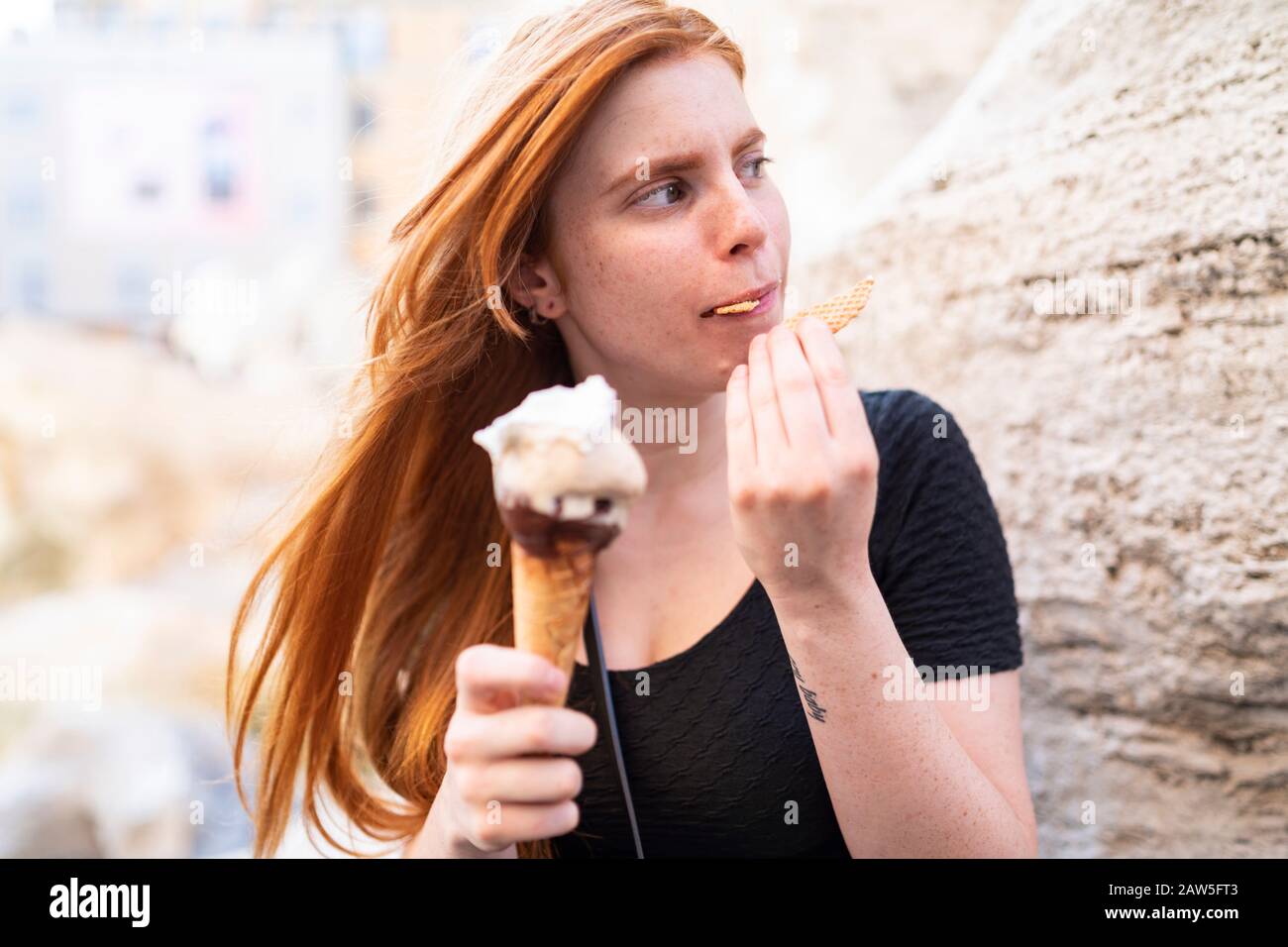 Hübsche, tausendjährige Frau mit langen roten Haaren, die leckeren Eiskegel mit Keks genießen und im Sommer auf der Straße in der Stadt sitzen Stockfoto