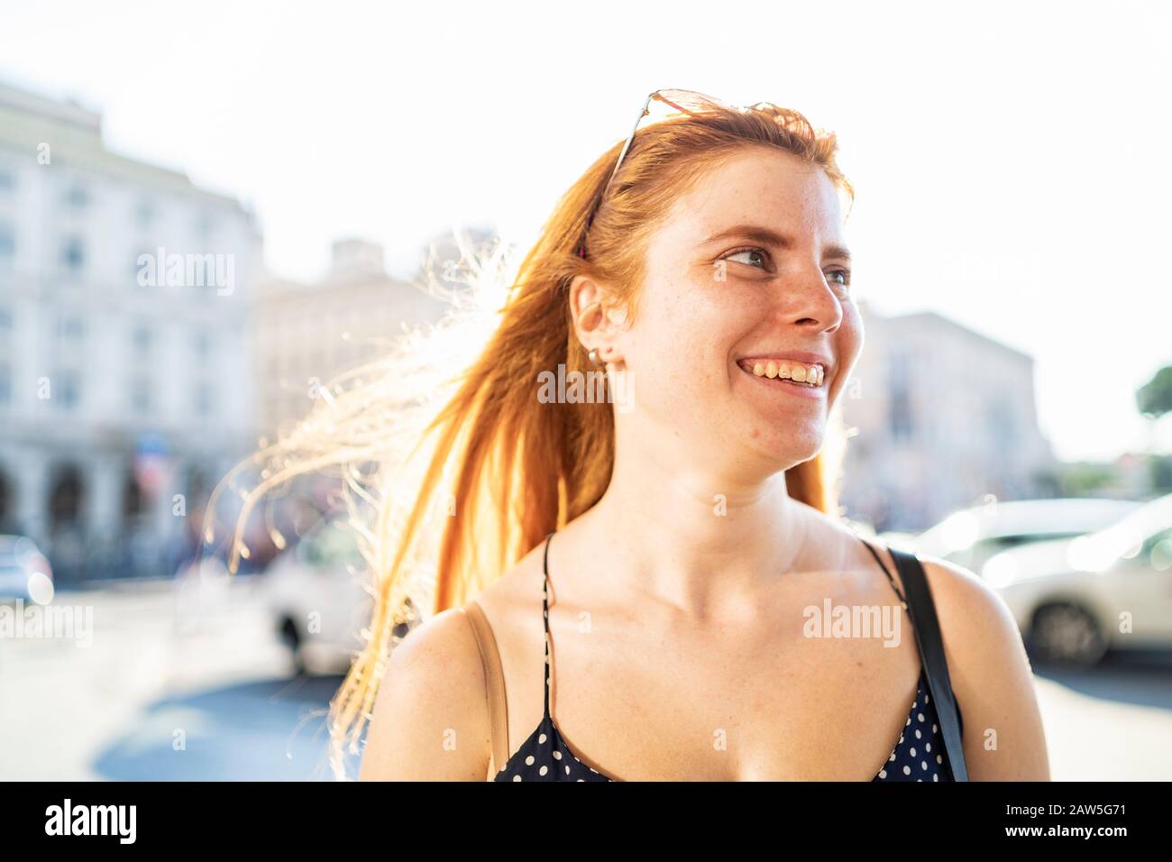 Glitzernde junge Frau mit rotem Haar lächelnd und mit Blick auf die Straße in der Stadt am sonnigen Sommertag Stockfoto