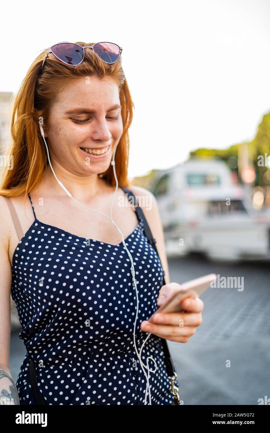 Glitzernde junge Frau mit Ingwerhaar mit Smartphone und Musik in Ohrhörern, während sie sich am sonnigen Sommertag auf der Straße ausruhen Stockfoto