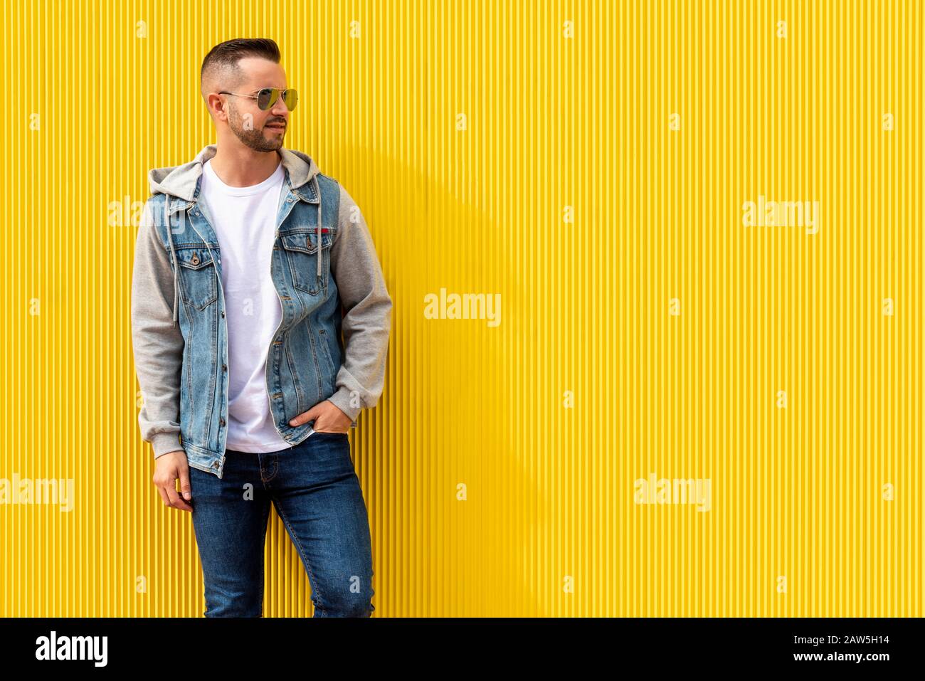 Porträt eines jungen, coolen Mannes über gelbem Hintergrund. Der Mann trägt eine blaue Jeans, eine Denimjacke und eine Sonnenbrille. Stockfoto
