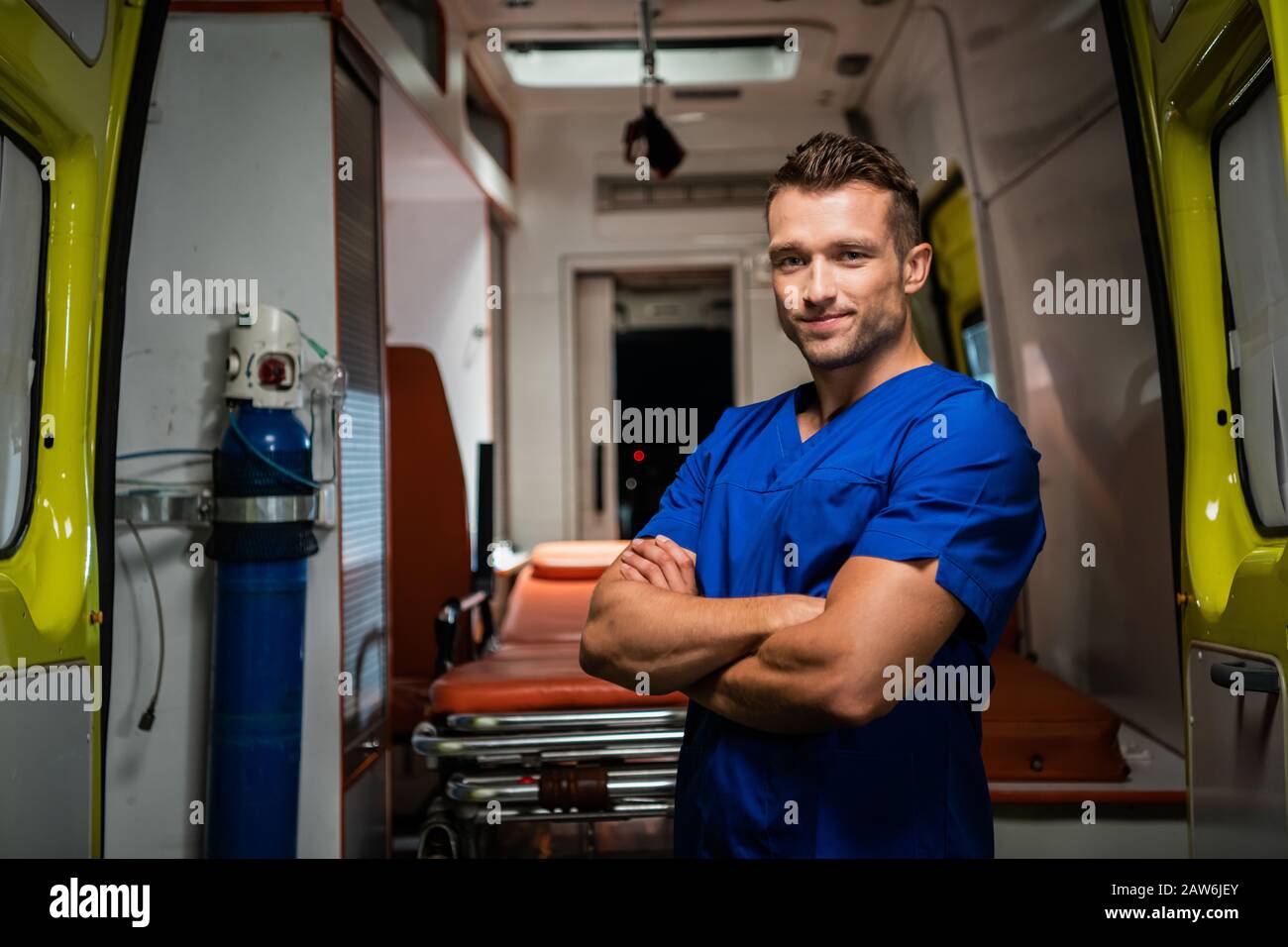 Junge glückliche Sanitäter in medizinischer Uniform sehen Kamera und Lächeln an, Rettungswagen auf dem Hintergrund Stockfoto