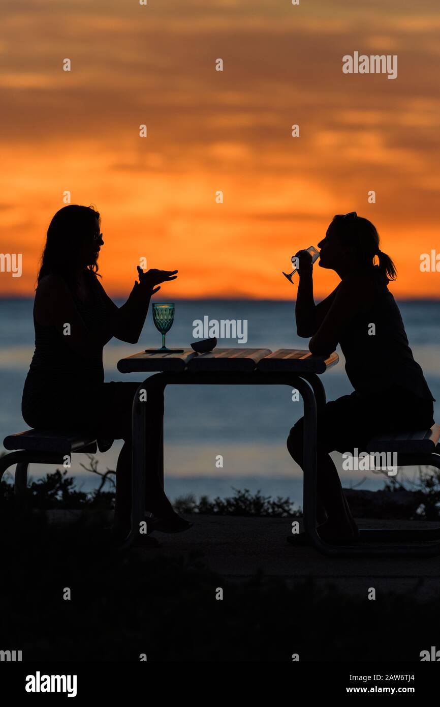 Eine leuchtend orangefarbene Silhouette bei Sonnenuntergang, die zwei Ökotouristen mit einem Happy Hour Drink über dem Indischen Ozean in der Osprey Bay, Cape Range National Park in WA, verzaubern lässt. Stockfoto