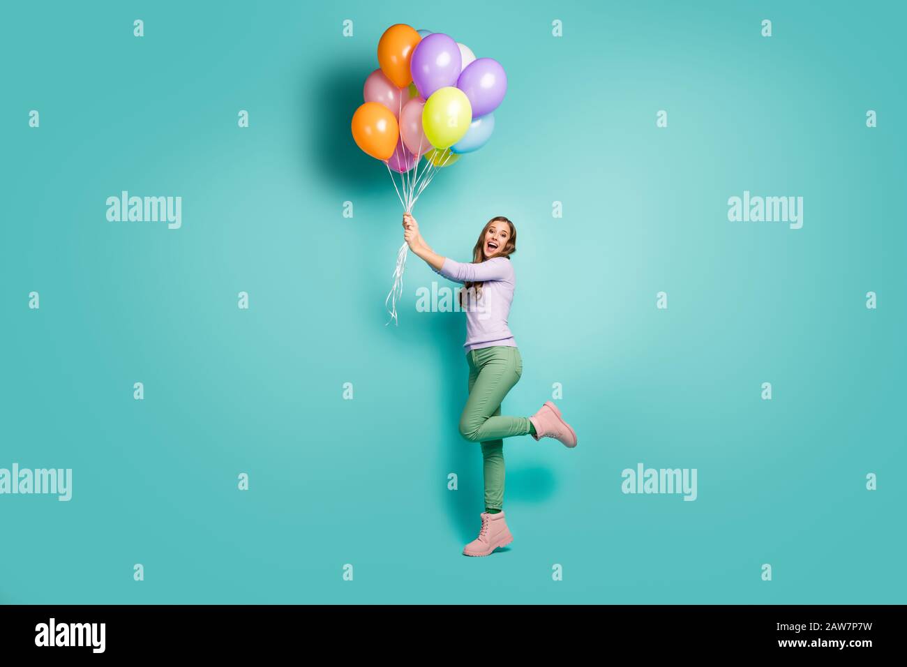 Das Profilfoto der hübschen funky Lady hält viele bunte Luftballons, die mit Wind wehenden, lilakischen Pullover, grünen Hosen, Stiefeln wegfliegen Stockfoto