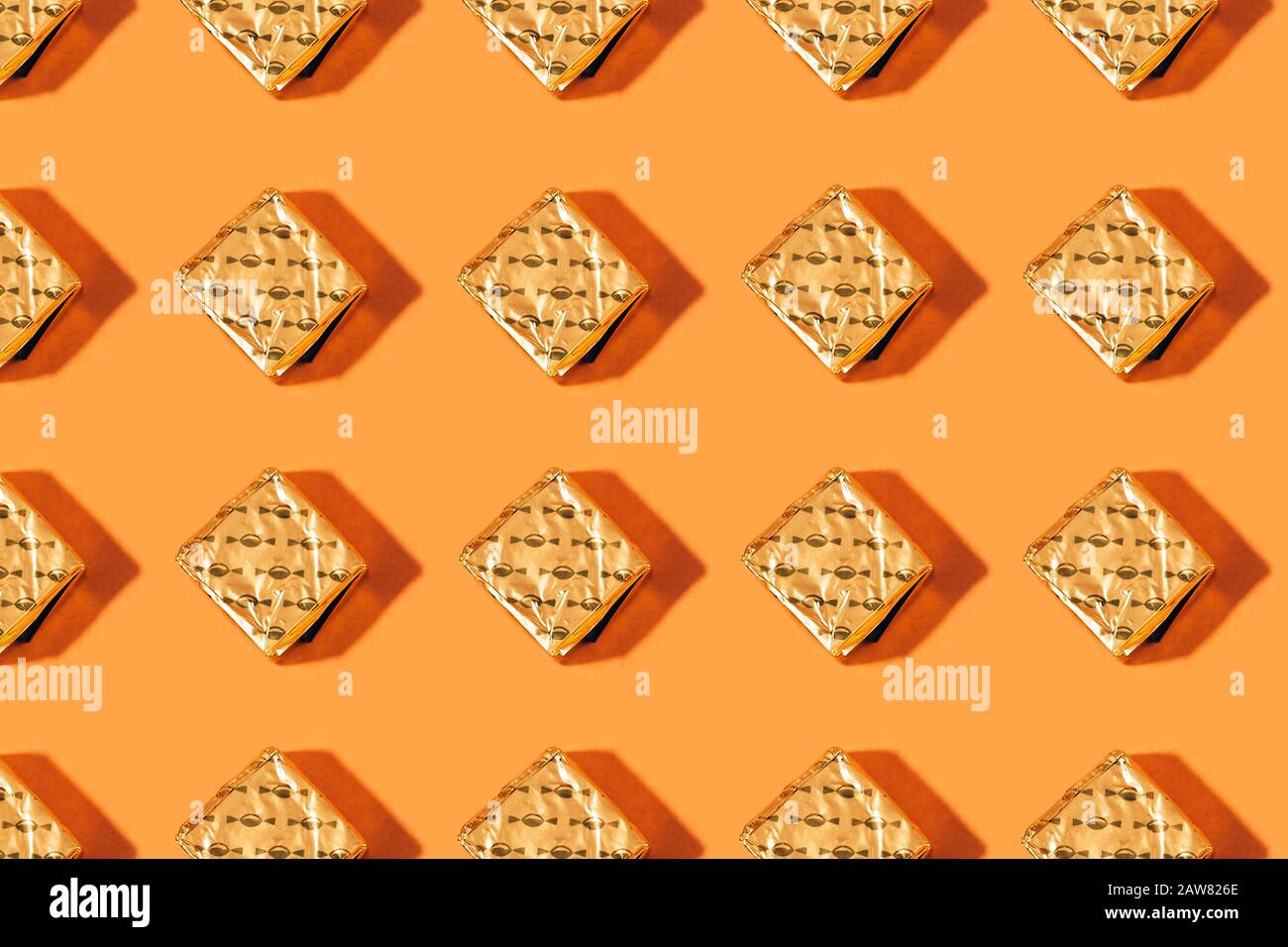 Pralinen wrappert goldfarben, auf orangefarbenem Hintergrund, sich wiederholendes Muster mit harten Schatten. Stockfoto