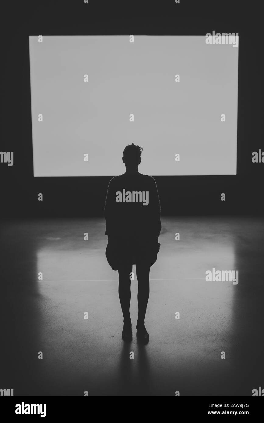 Dramatisches Schwarz-Weiß-Bild einer Silhouette einer menschlichen weiblichen Figur, die mit einem beleuchteten Bildschirm im Hintergrund auf dem Rücken zur Kamera steht Stockfoto