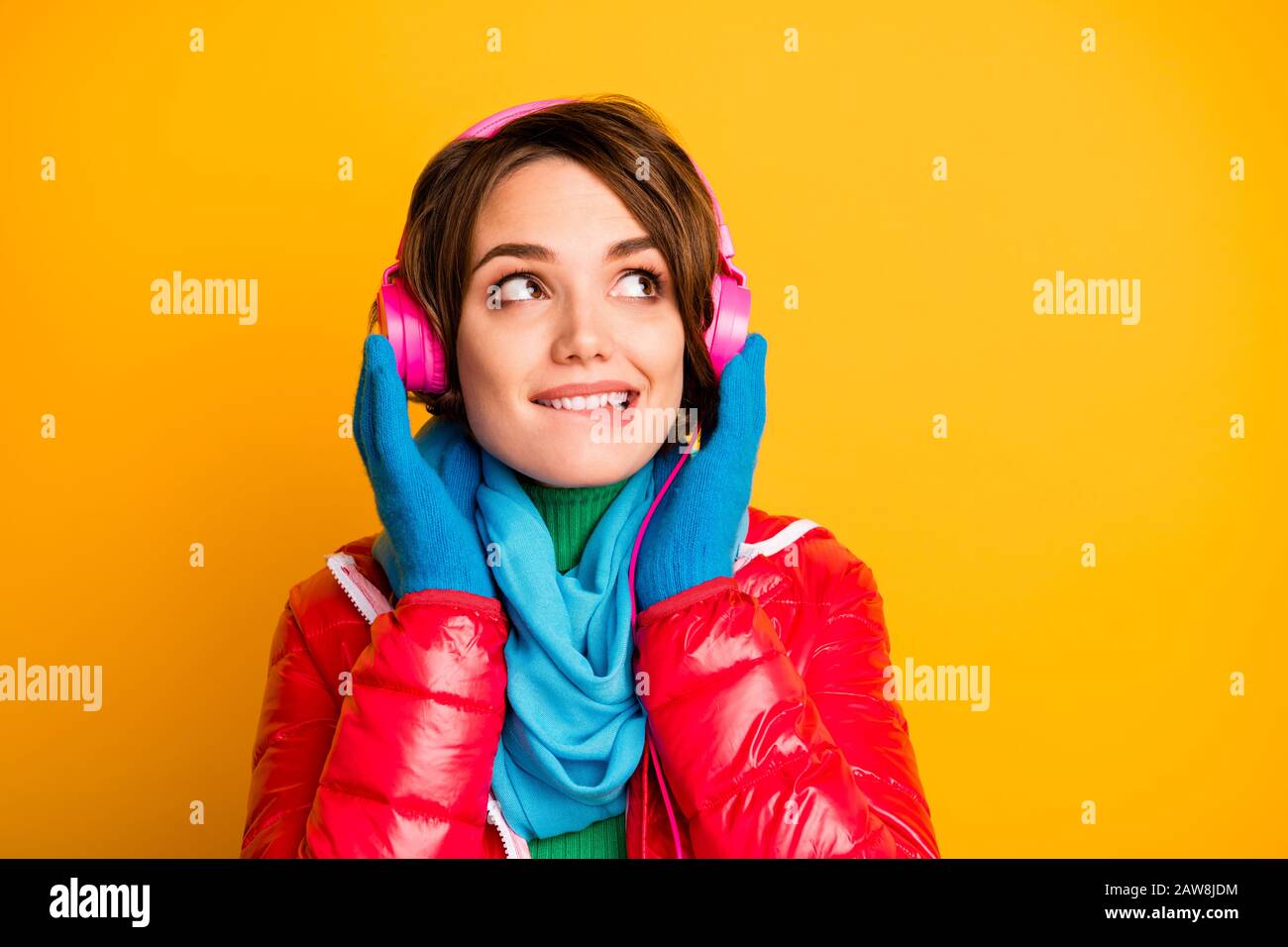 Closeup-Foto von hübscher Dame beißende Lippen hören Sie moderne Kopfhörer mit leerem Raum und stilvollem rotem Mantel blaue Schal Handschuhe Stockfoto