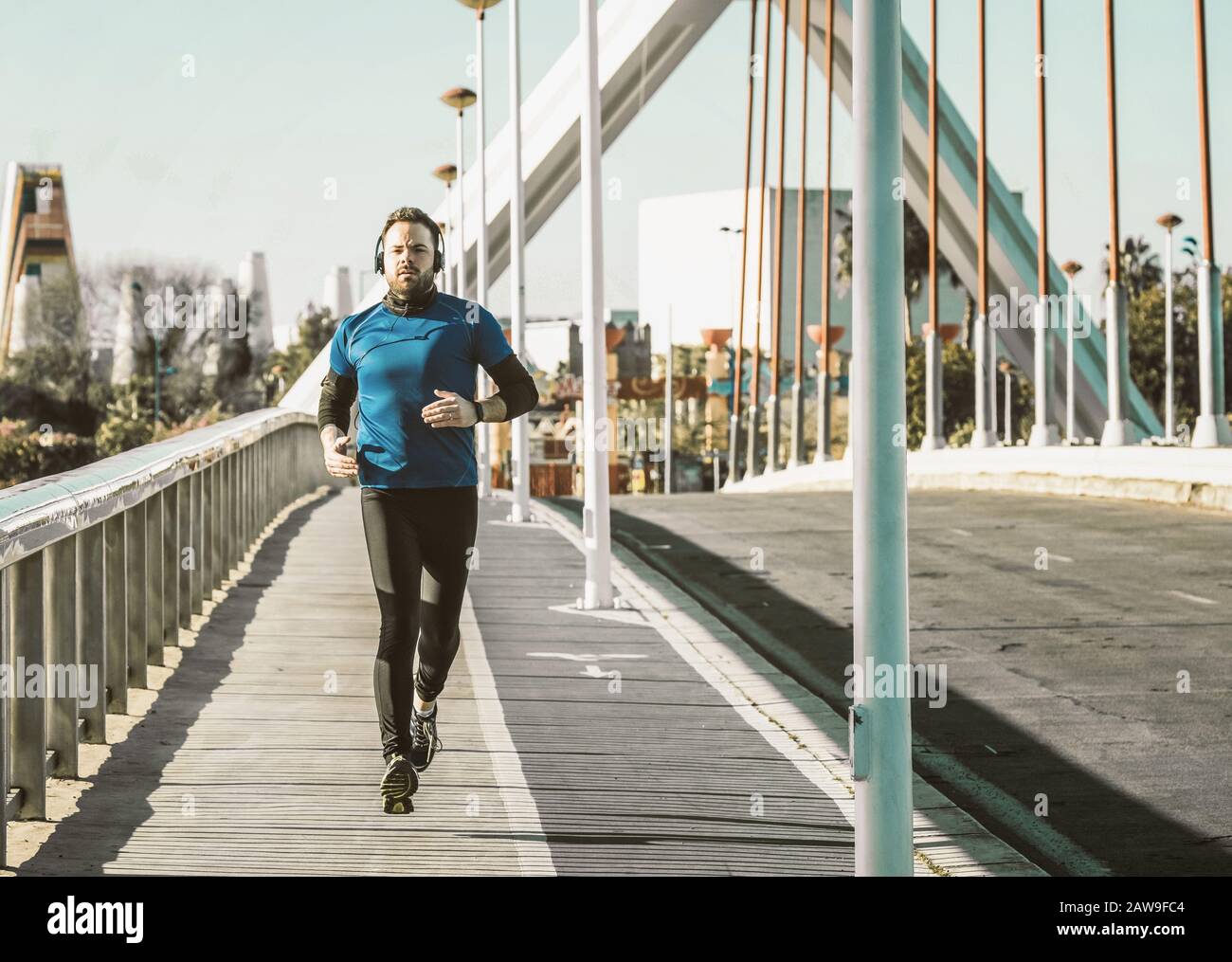 Junger Mann, der auf einer Brücke in einem blauen Hemd läuft und mit einem Paar Kopfhörern Musik hört. Stockfoto