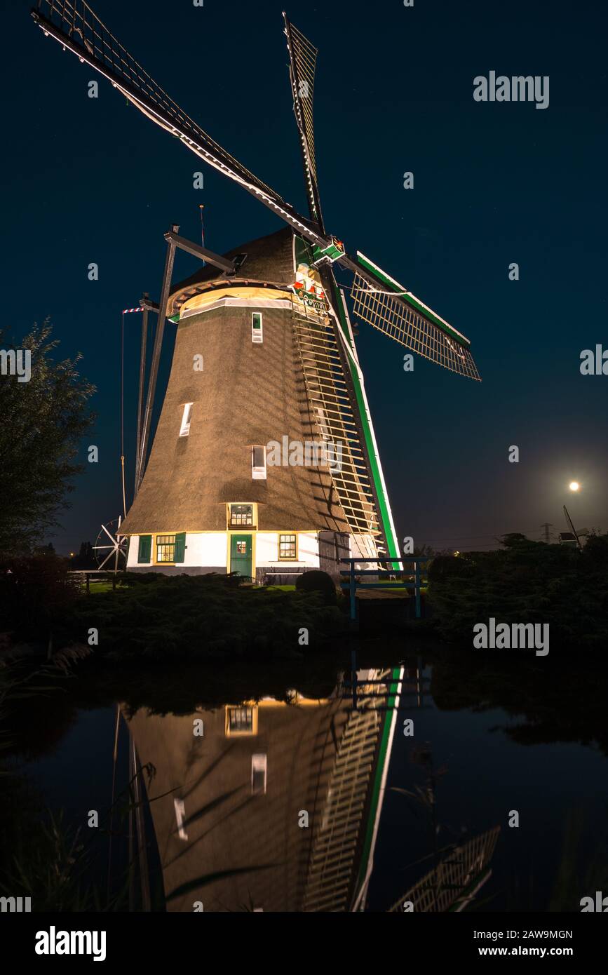 Die klassische holländerwindmühle wird nachts mit schönen Lichtreflexionen im Wasser beleuchtet. Steigender Vollmond im Hintergrund. Stockfoto