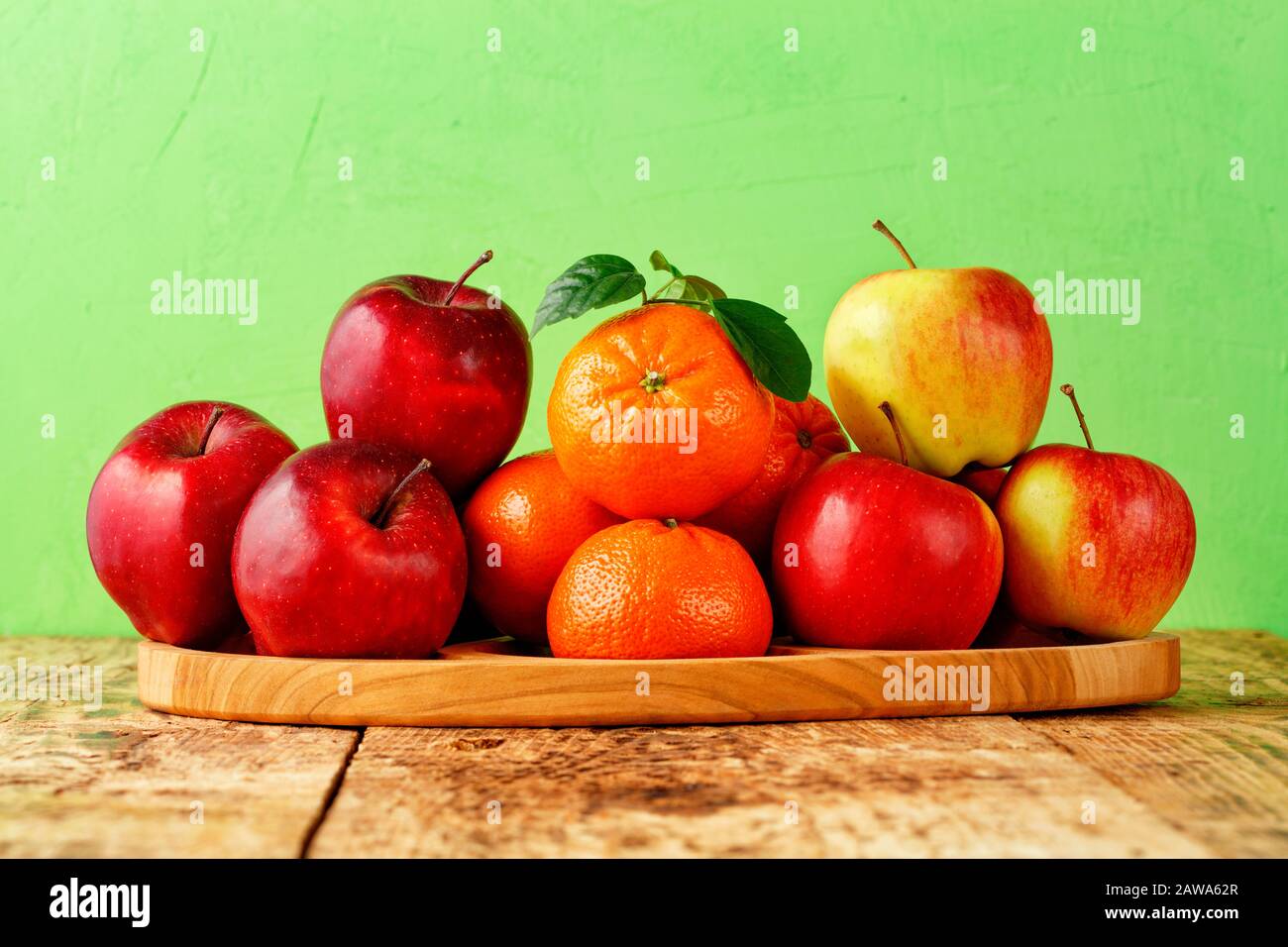 Rote reife Äpfel und Mandarinen mit grünen Blättern liegen auf einem Holztablett auf einem alten Holztisch auf hellgrünem Hintergrund, Bild mit Kopierraum. Stockfoto