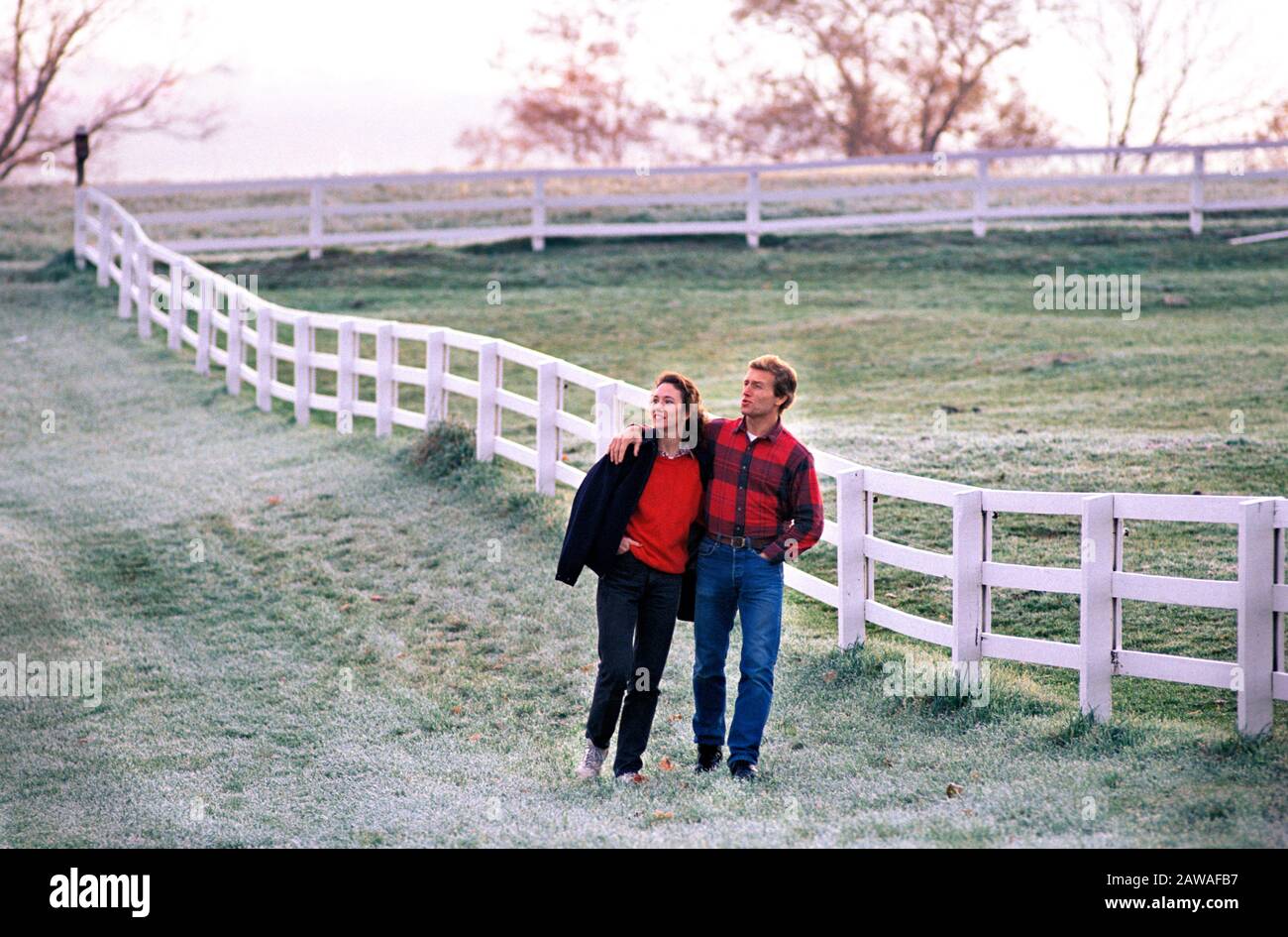 Kaukasisches Paar, das auf dem mit Tau bedeckten Feld auf dem Bauernhof entlang des Zauns läuft Stockfoto