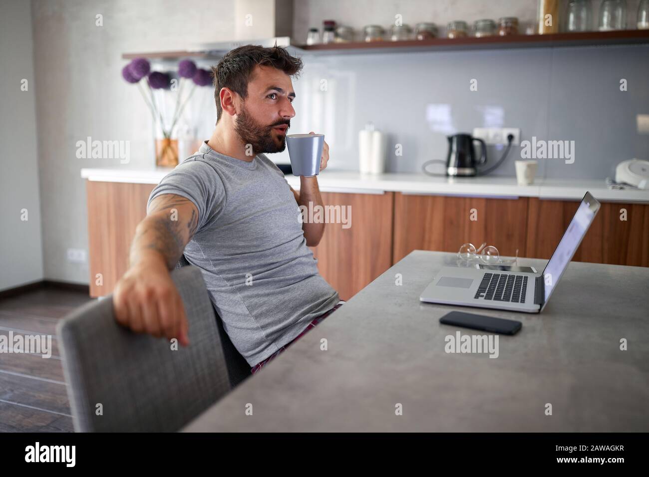 Junger Erwachsener mit Bartsitzend und trinkendem Kaffee vor dem Laptop auf dem Tisch. Moderner Lebensstil, entspanntes, geschäftliches Konzept Stockfoto