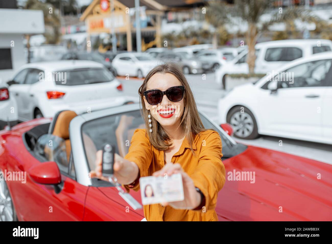 Porträt einer schönen jungen Frau, die mit Führerschein und Schlüssel in der Nähe des roten Cabriolets auf dem Parkplatz steht. Konzept eines glücklichen Kauf oder Miete eines Autos Stockfoto