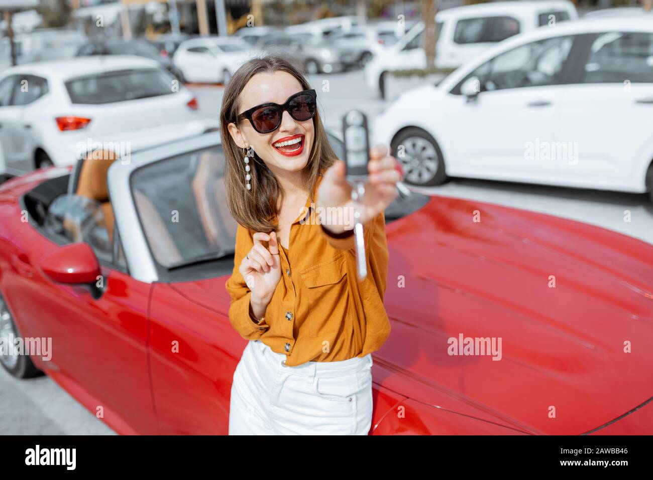 Porträt einer schönen jungen Frau, die mit Schlüsseln in der Nähe des roten Cabriolets auf dem Parkplatz steht. Konzept eines glücklichen Autokaufs oder -Mietens Stockfoto