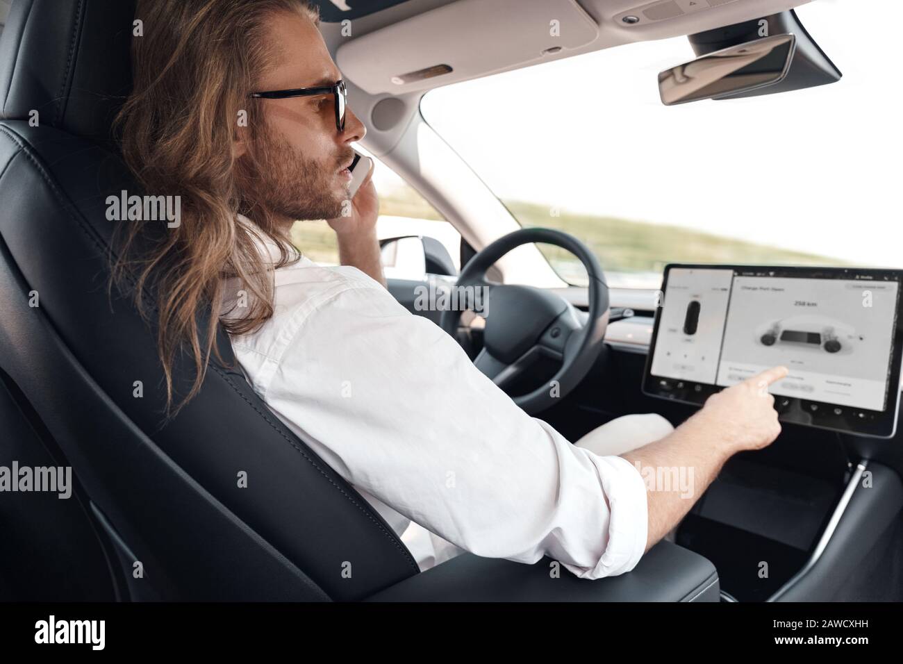 Transportmodus. Bärtiger Mann mit langen Haaren in der Sonnenbrille, der im Elektroauto sitzt und auf dem Autopiloten unterwegs ist. Er spricht über Smartphone-Touchign Stockfoto