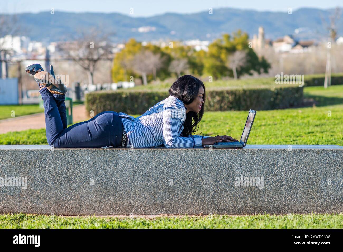Lächelnde afro-amerikanische Frau, die an einem sonnigen Tag mit einem Laptop in einem Außenpark auf einer Bank liegt. Stockfoto