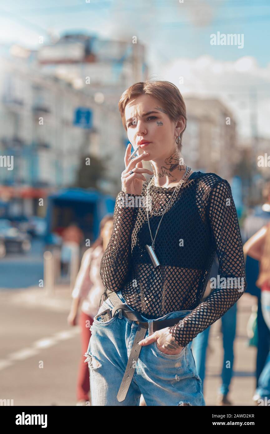 Urbaner Stil. Junge, stilvolle Frau, die im Freien Hand in der Tasche steht, raucht eine Zigarette, die nachdenklich beiseite schaut Stockfoto