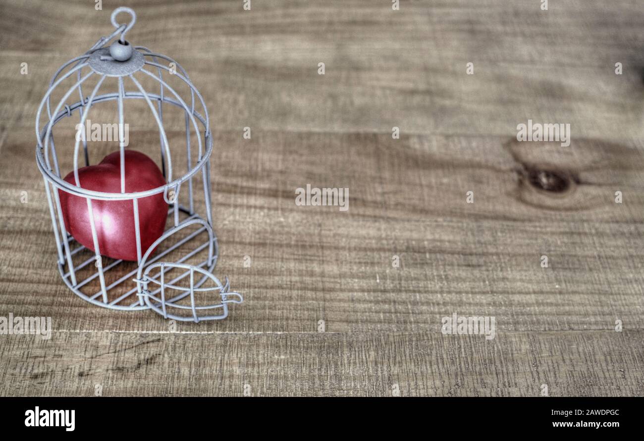 Rotes Herz in einem Vogelkäfig auf einem Holztisch. Selektive Fokussierung auf die offene Tür. Kopierbereich. Stockfoto