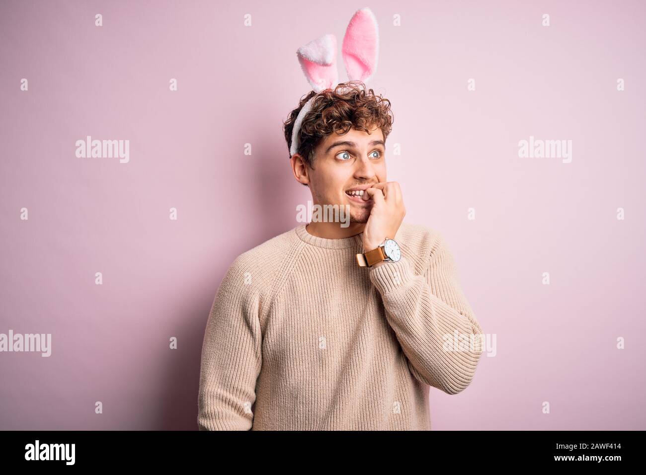 Junger, gutaussehender blonder Mann, der easter Kaninchenohren über isoliertem rosafarbenem Hintergrund trägt und gestresst und nervös aussieht, während die Hände an den Mundnägeln beißen. Anxie Stockfoto