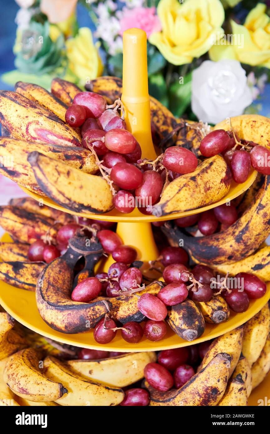 Nahaufnahme von überreifen gefleckten Bananen und roten Trauben auf einem Kunststoff-Turmblech mit bunten Plastikblumen im Hintergrund Stockfoto