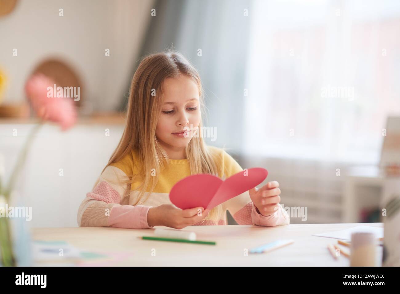 Portrait des niedlichen kleinen Mädchens, das herzförmige Karte hält und lächelt, während es am Tisch im gemütlichen Innenbereich sitzt, Platz für Kopien Stockfoto
