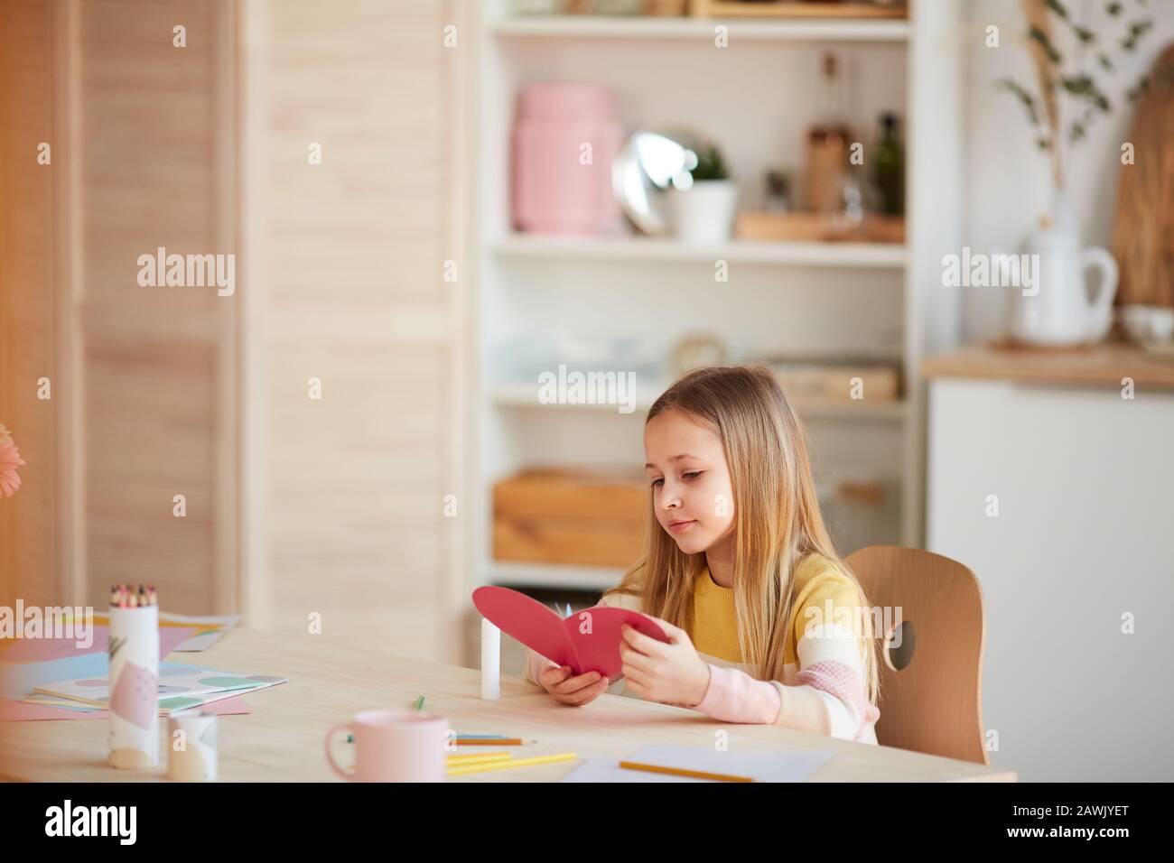 Warmes Porträt eines süßen Mädchens, das eine herzförmige Karte macht und lächelt, während es im gemütlichen Zuhause am Tisch sitzt und Platz für Kopien bietet Stockfoto