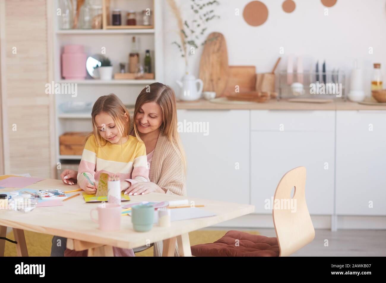 Warmtoniges Porträt der glücklichen Mutter, die kleine Tochter umarmt, während sie Bilder auf Holztisch in der Küche malte, Kopierraum Stockfoto
