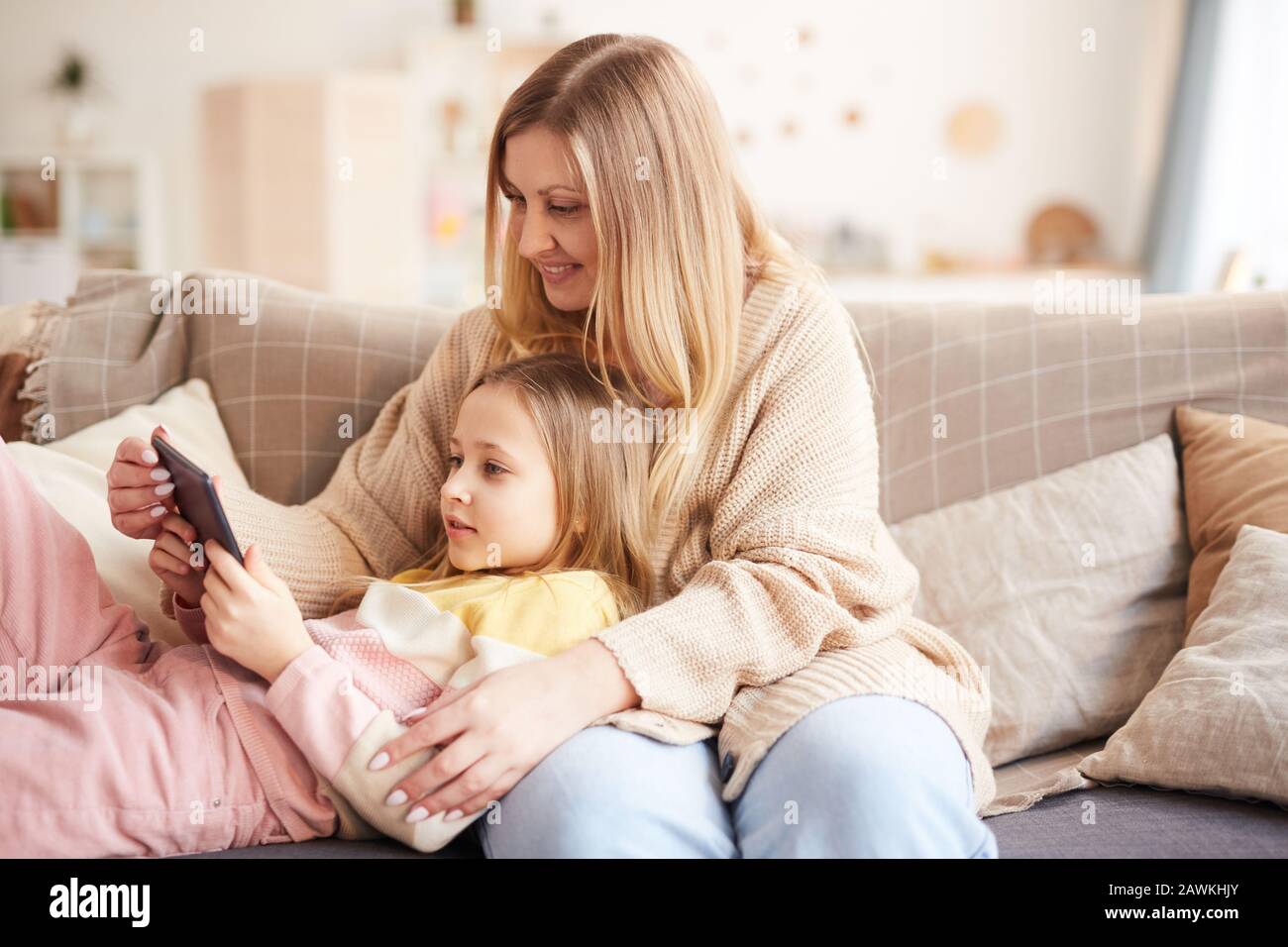 Warmes Porträt der modernen Mutter, die mit niedlichem Mädchen spielt, während sie die Zeit auf der Couch genießen und digitale Tablette und Kopierer verwenden Stockfoto
