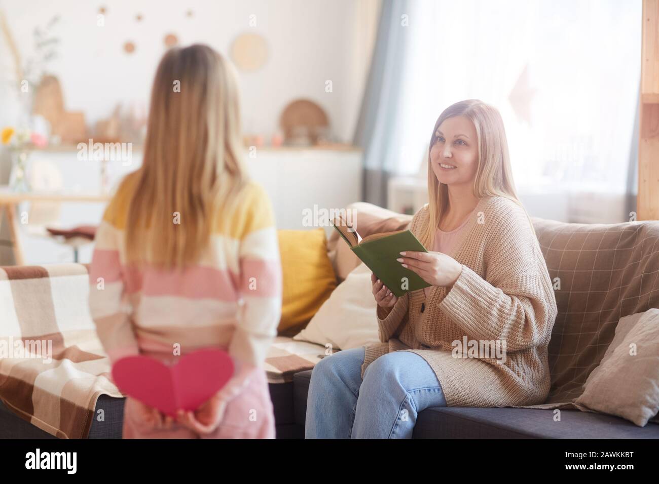 Warmtoniges Porträt der fröhlichen modernen Mutter, die Tochter betrachtet, die sie am Muttertag überrascht, während sie Buch auf der Couch in gemütlichem Innenleben liest, Kopierer Stockfoto