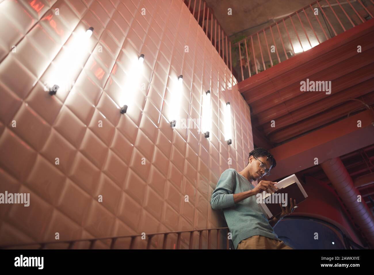 Weitwinkel-Porträt des jungen afroamerikanischen Mannes, der Bücher hält, während er sich an die Wand im College-Campus lehnt, Copy-Space Stockfoto
