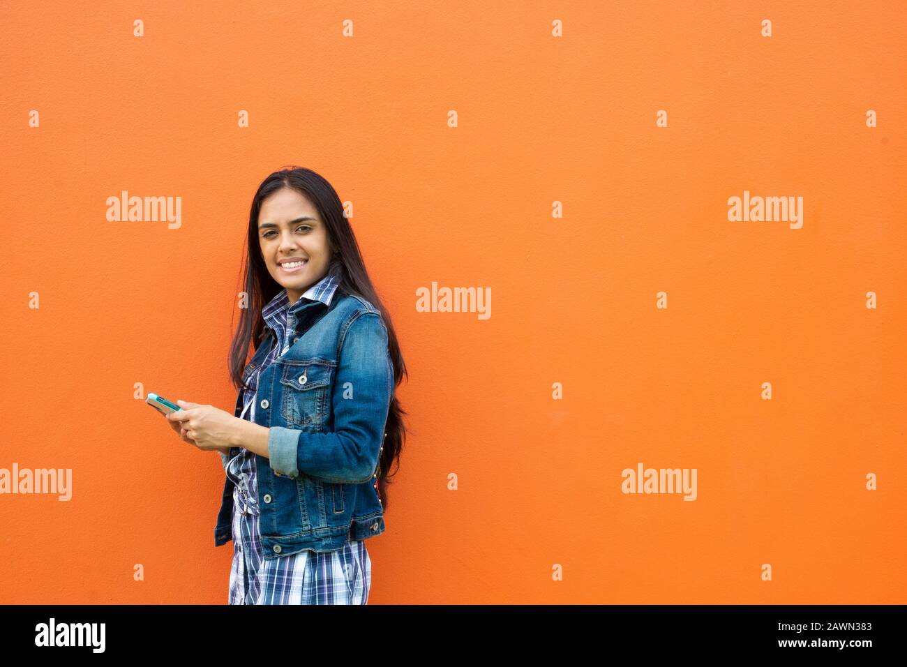Porträt Einer Lächelnden Jungen Frau mit Handy, Während sie Gegen Orange Wall, Panama, Mittelamerika steht Stockfoto