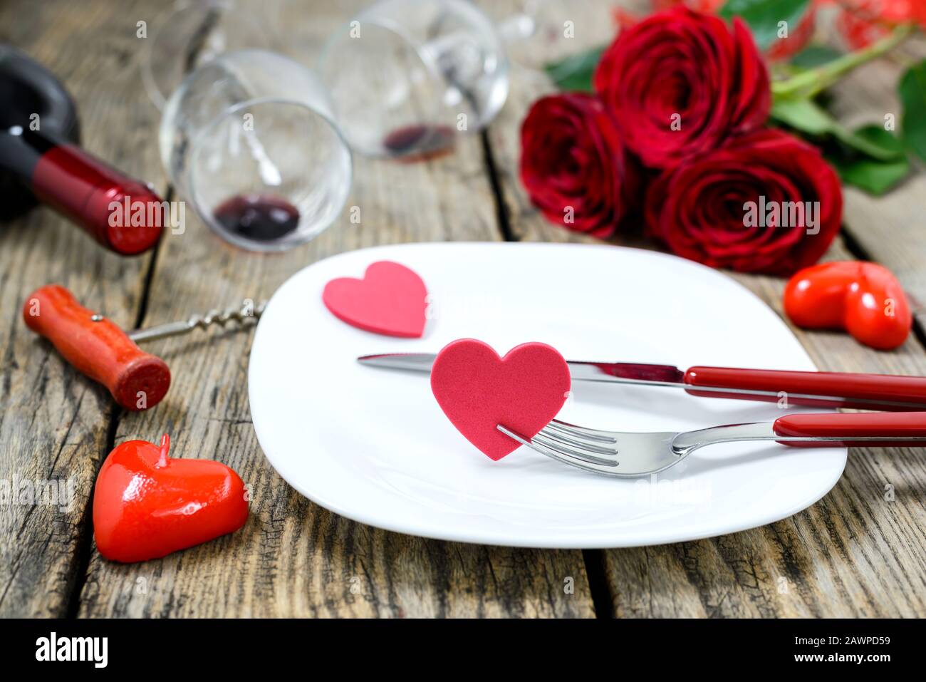 Herzen auf einem Teller mit а Gabel und Messer, а Weinflasche, Korkenzieher, zwei Weinglas, rote Rosen und Kerzen auf einem alten Holztisch. Selektiver Fokus. Stockfoto