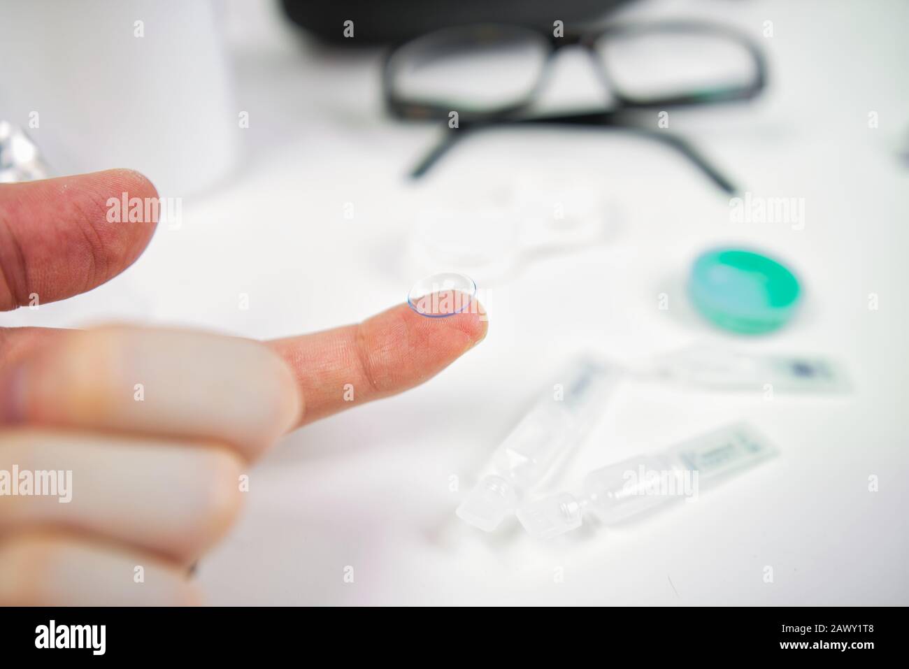 Hand mit Kontaktlinse, die in die Augen eingesetzt werden kann. Im Hintergrund sichtbare Brille, Tropfen und Hüllen Stockfoto
