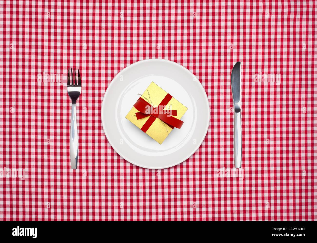 Überraschungsmahlzeit, ein Geschenk verpackt auf einem runden weißen Teller mit Messer und Gabel von oben auf einem roten Gingham Tischtuch Stockfoto