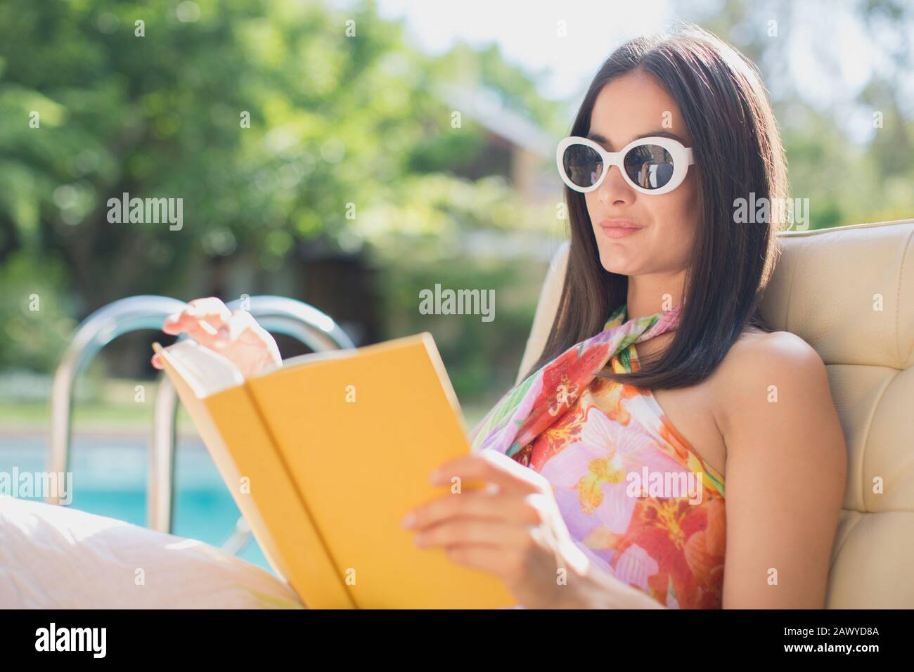 Frau in Sonnenbrille liest Buch im sonnigen Sommer am Pool Stockfoto