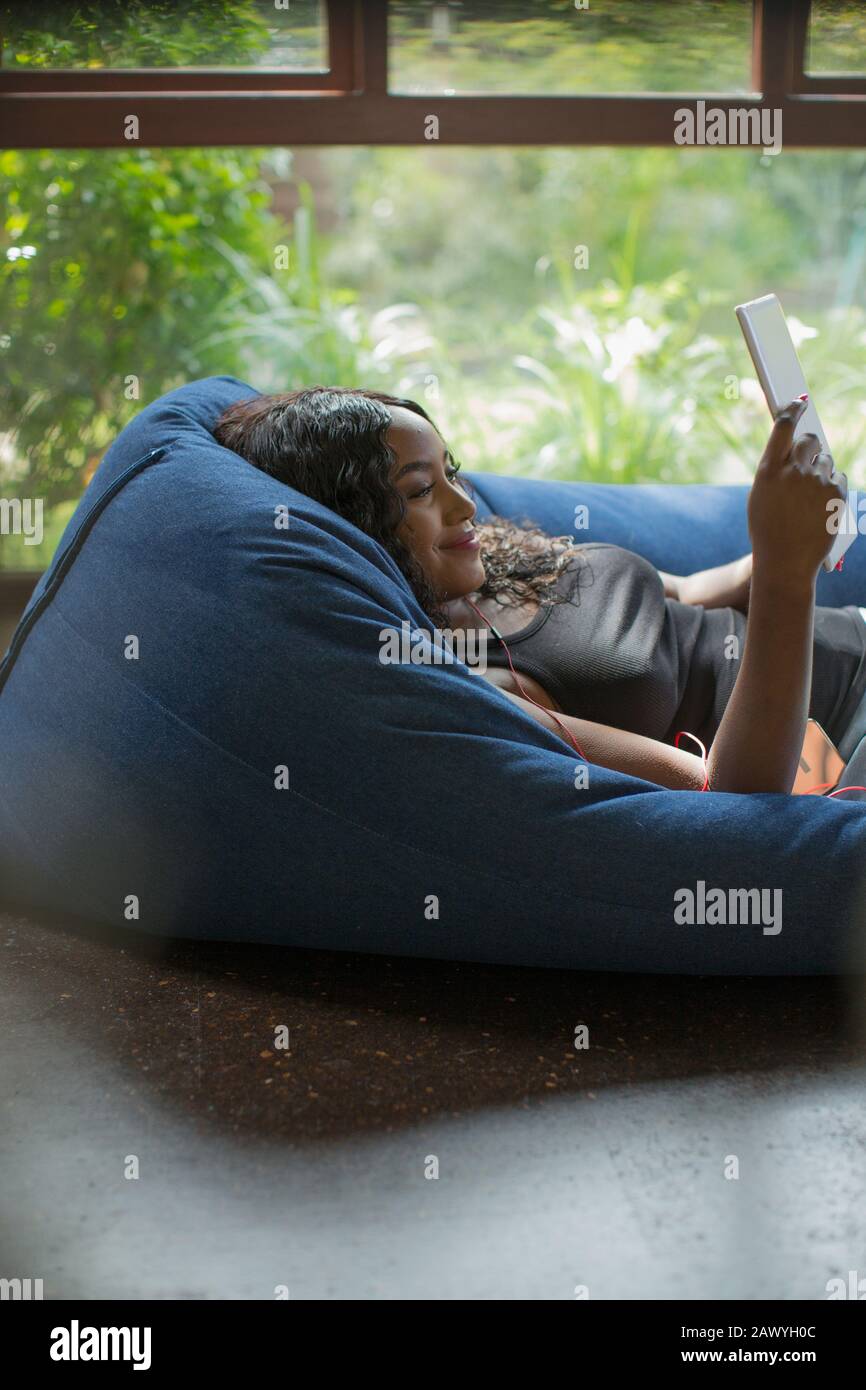 Fröhliche junge Frau, die sich mit einem digitalen Tablett im Beansack-Stuhl entspannt Stockfoto