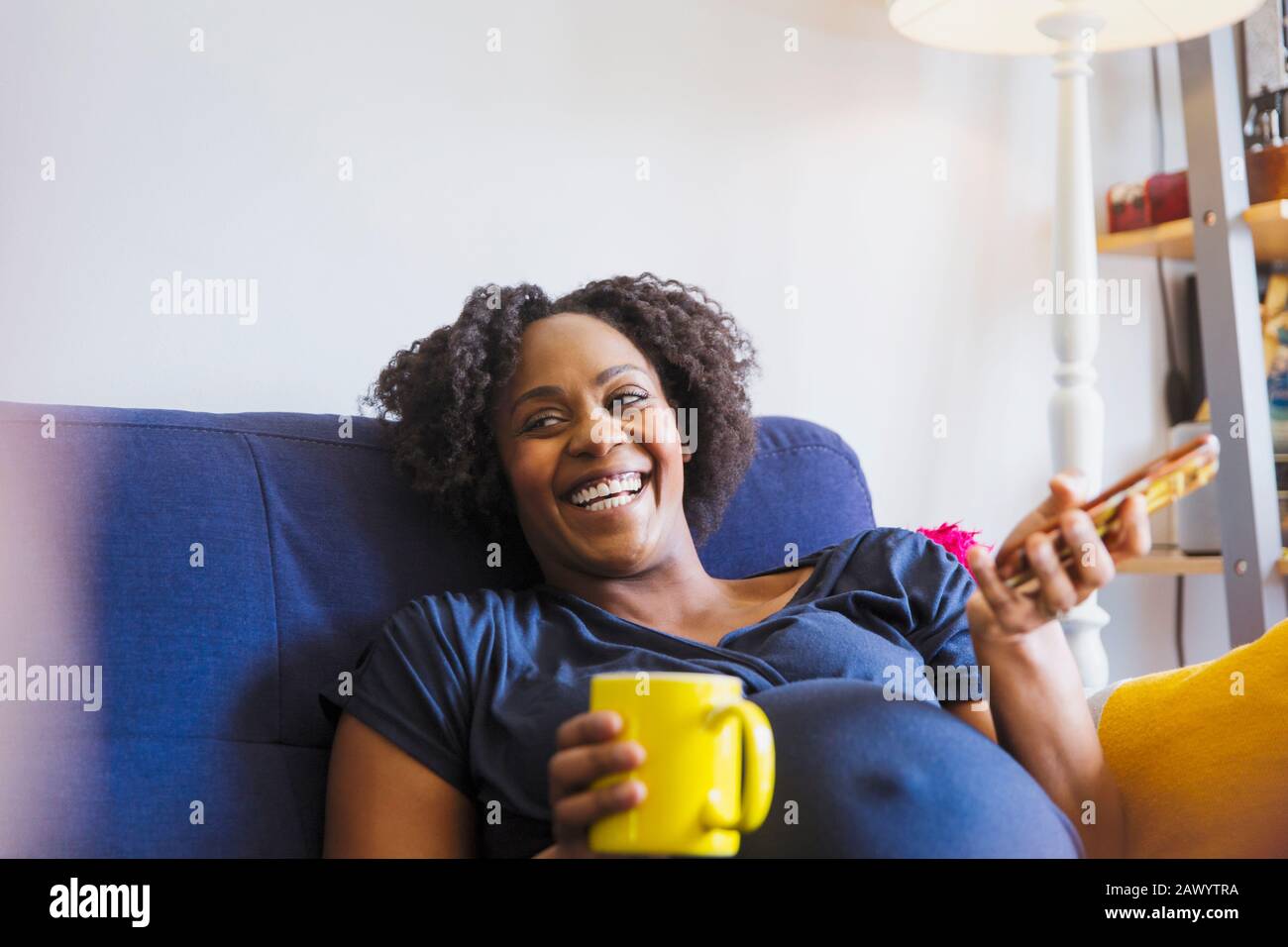 Schwangere Frau lachen mit Tee und Smartphone auf dem Sofa Stockfoto