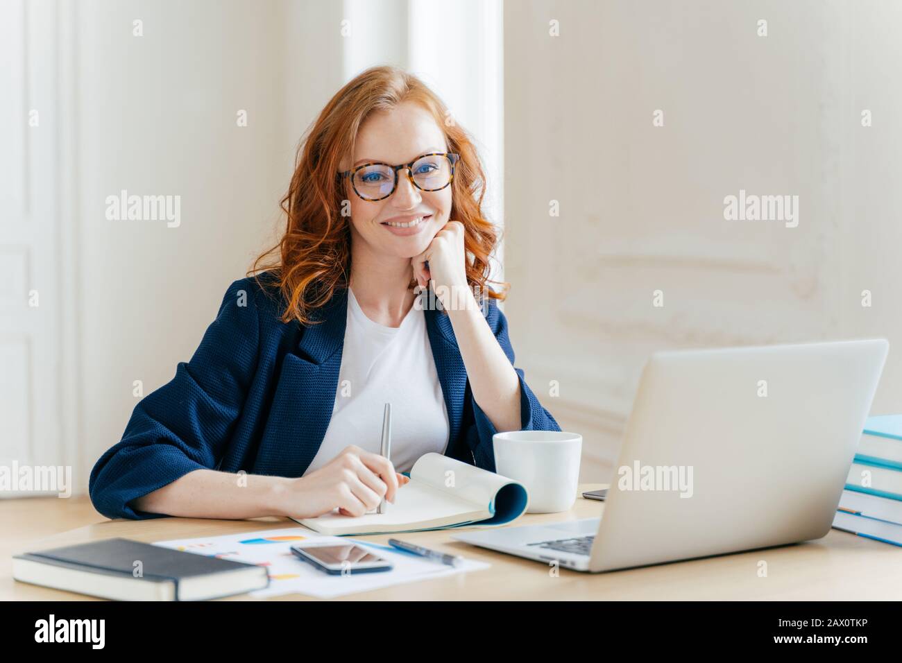 Erfolgreicher Ökonom in Brille, überprüft Informationen auf dem Laptop, liest Informationen auf der Finanzwebsite, schreibt in Notizblock und trinkt heißen Kaffee Stockfoto