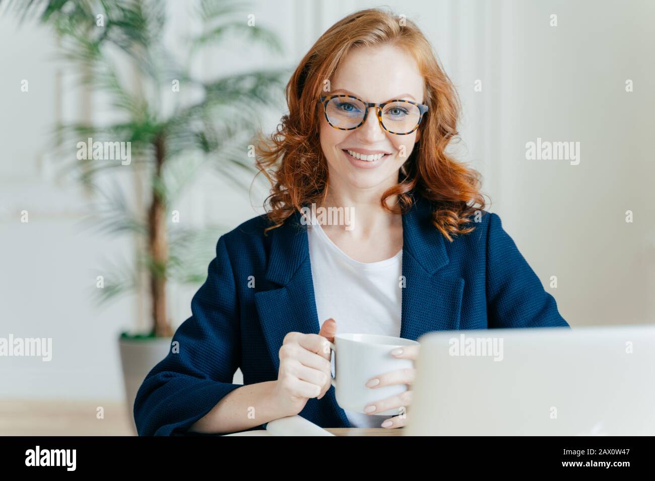 Fröhliche Rotkopffrau bereitet Bewerbungsschreiben vor, sitzt vor geöffnetem Laptop, durchsucht das Internet und sucht nach Stellenangeboten, hält MUG w Stockfoto