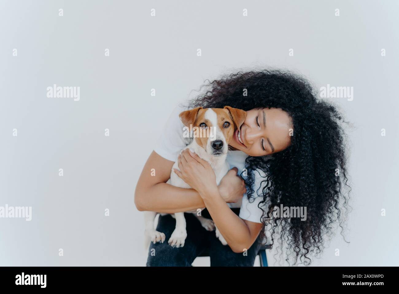 Bild von verspielter hübscher Frau mit Afro Frisur, lächelt freudig, hält Haustier, trägt Hund zu veterian, isoliert über weißem Hintergrund. B. Stockfoto