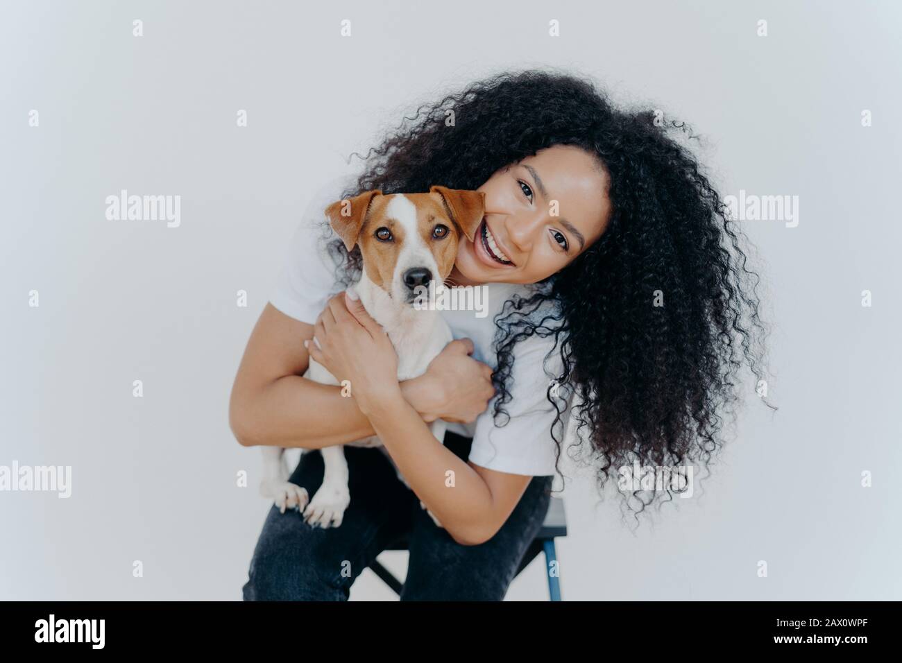 Fröhliche lockige Frau neigt den Kopf, hält Stammhund, hat fröhlichen Ausdruck, lächelt angenehm, hat lockige Frisur, trägt weißes T-Shirt, isoliert. Afro Stockfoto