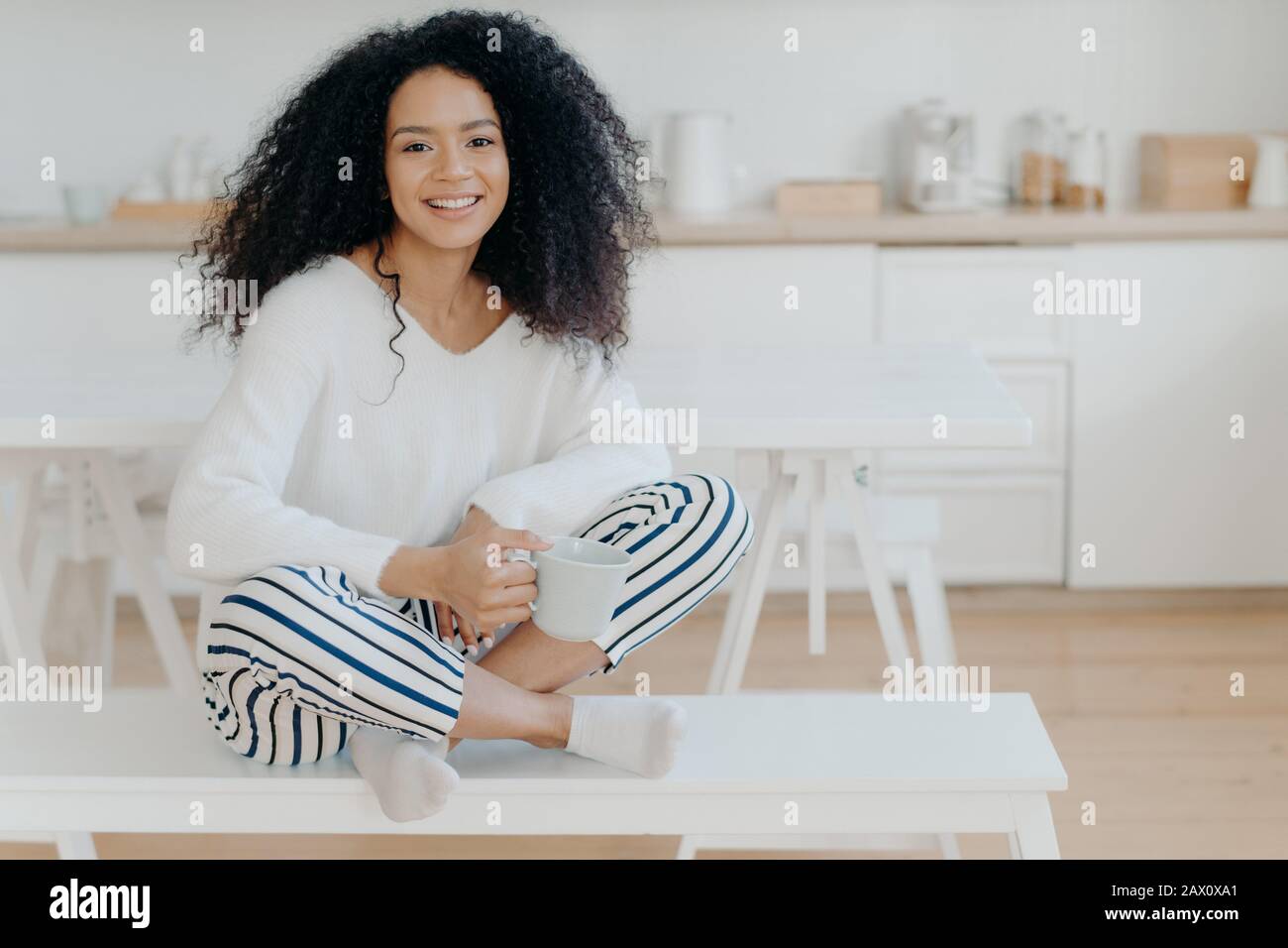 Lächelnde, entspannte afroamerikanische Frau sitzt gekreuzte Beine auf der Bank gegen den Kücheninnenraum, trägt weißen Pullover und gestreifte Hosen, trinkt heißen Beverag Stockfoto