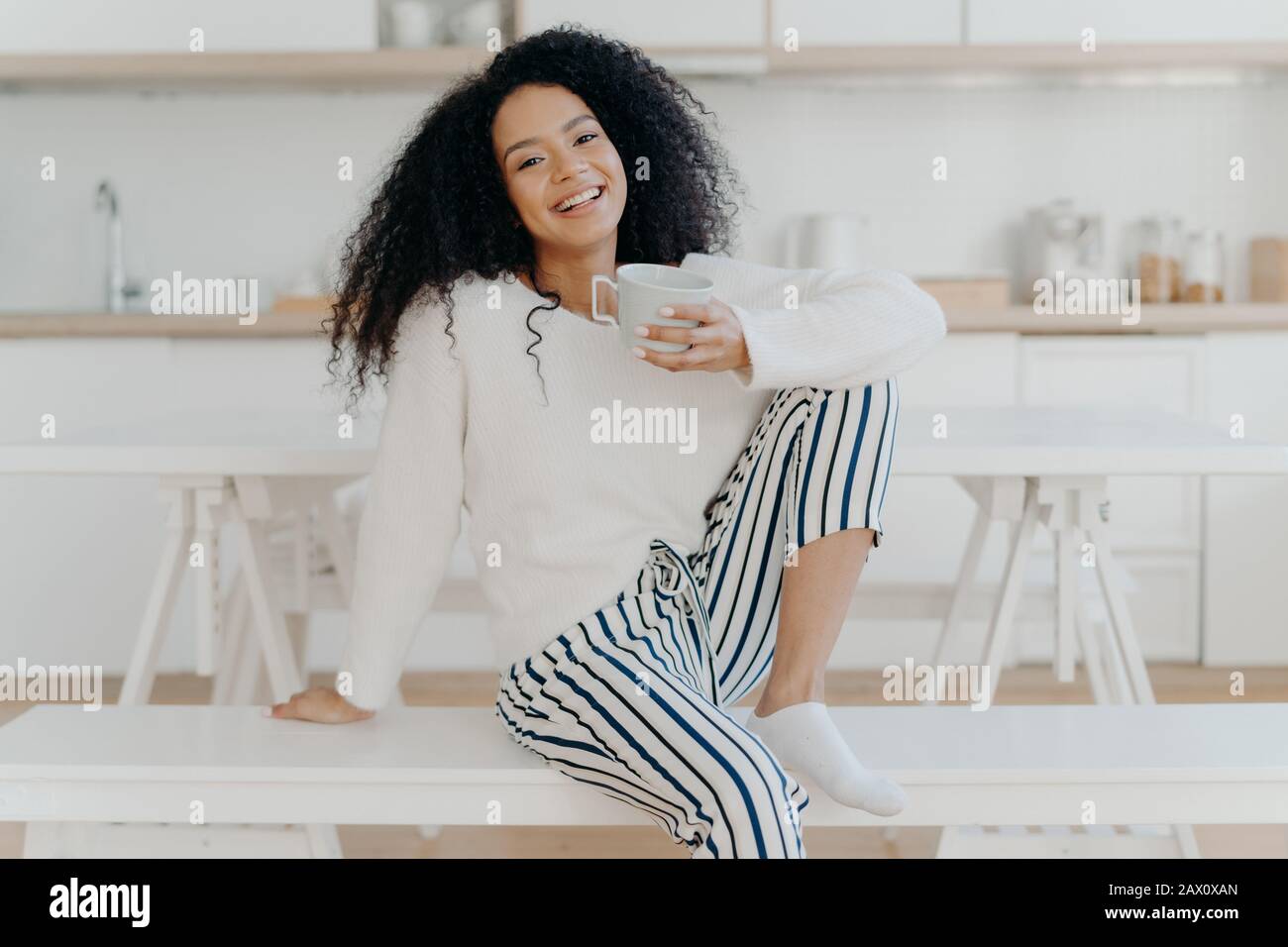 Schöne junge Frau mit Afro-Haarschnitt, posiert auf weißer Bank im Innenbereich, in stilvoller Kleidung gekleidet, genießt aromatisches Getränk, genießt zu Hause Frühstück Stockfoto