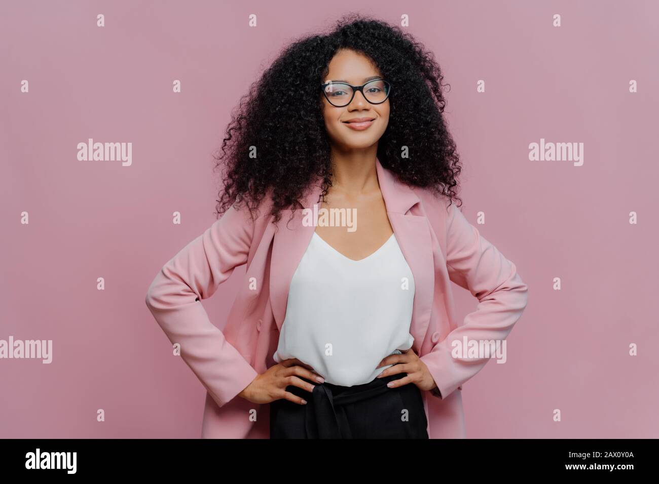Taille up Schuss von zufriedenen dunkelhäutigen Büroangestellten hält die Hände auf ihrer schlanken Figur, hat einen fröhlichen Look, trägt transparente Brille, posiert über pur Stockfoto