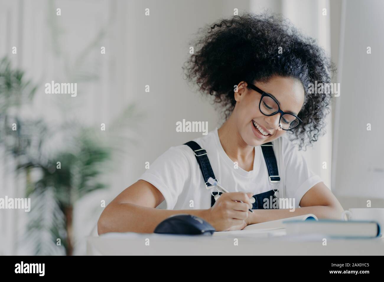 Die fröhliche afro-amerikanische Studentin schreibt Informationen auf, bereitet sich auf Prüfungen vor, sitzt im feigen Raum, hat geschweifte dunkle Haare, trägt eine optische Brille WH Stockfoto