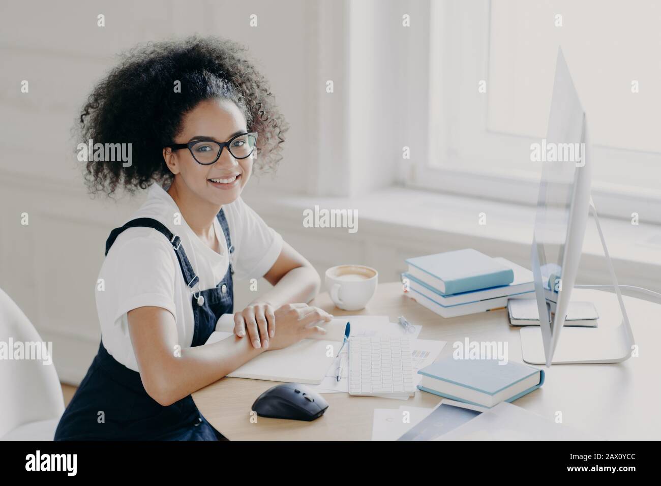 Positive afro-amerikanische Freiberuflerin posiert am Arbeitsplatz mit Papieren und Lehrbüchern, arbeitet ferngesteuert am Computer, hat Kaffeepause, arbeitet selbst Stockfoto