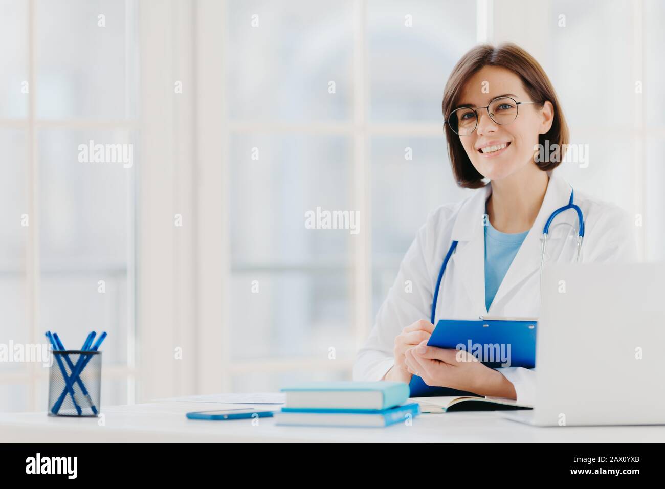 Der weibliche Arzt schreibt das Rezept auf Sonderform, arbeitet in der Privatklinik, trägt einen weißen medizinischen Gown, der bereit ist, Patienten zu sehen, Posen am Arbeitsplatz. S Stockfoto