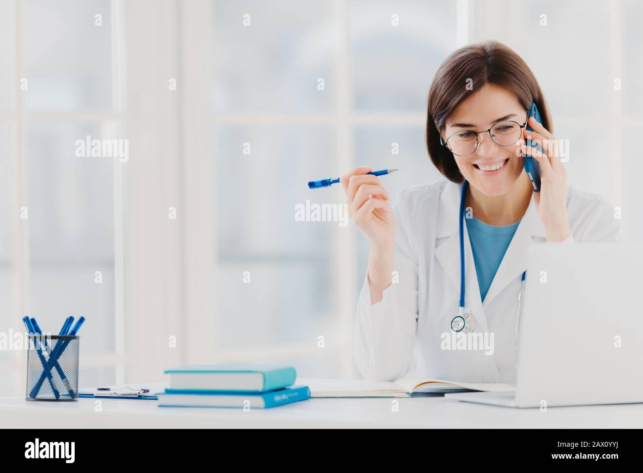 Der froh-professionelle Arzt, der sich auf den modernen Laptop konzentriert, nützliche Informationen liest, telefonische Gespräche führt, medizinische Probleme diskutiert, sitzt Stockfoto