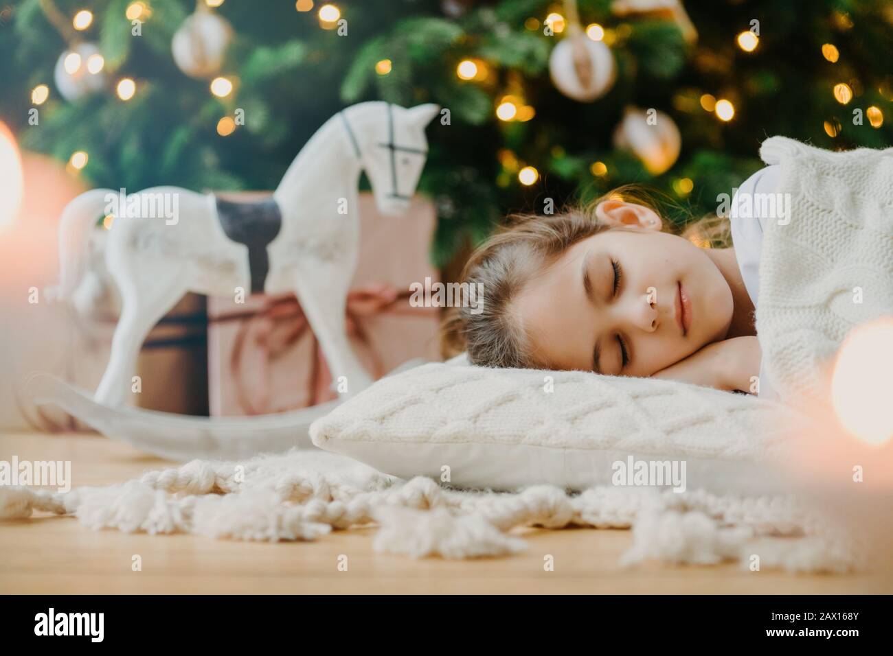 Horizontaler Schuss von entspannten Mädchen mit weißem Pullover, hat angenehme Träume, liegt unter dekoriertem Weihnachtsbaum, Spielzeugrocker und Geschenkschachteln herum Stockfoto